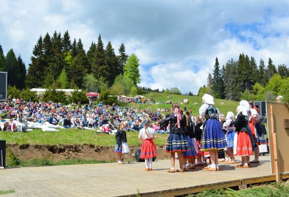 V popredí pódium s deťmi v krojoch, v pozadí ľudia sediaci na lúke na Poľane V popredí pódium s deťmi v krojoch, v pozadí ľudia sediaci na lúke na Poľane
