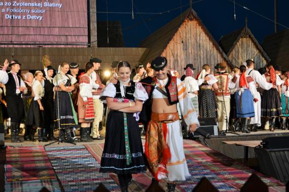Muž a žena v kroji v popredí, ďalší súboristi na pódiu folklórnych slávností. Muž a žena v kroji v popredí, ďalší súboristi na pódiu folklórnych slávností.